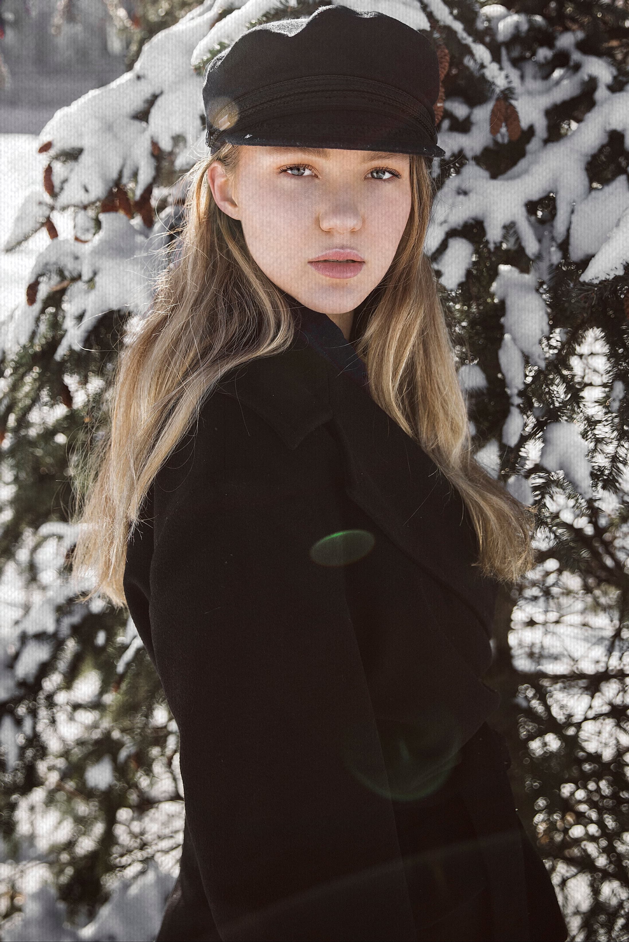 Portrait of a woman wearing a black baker boy hat and a black coat standing in front of a snowy pine tree.