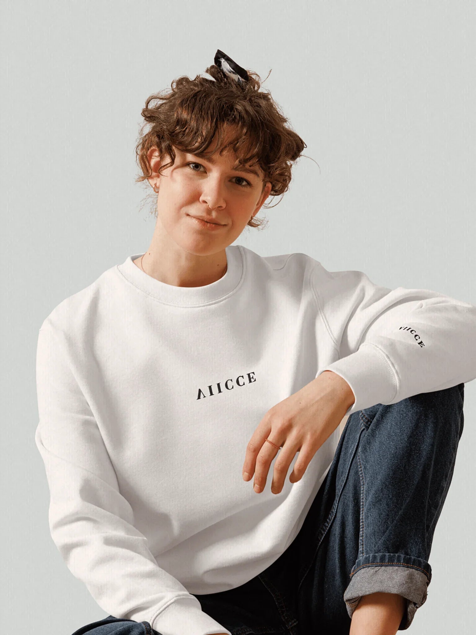 Signature Box Sweatshirt | Ash White. Female model sitting front-facing with the oversized sweatshirt featuring a vertically inverted embroidered logo on the chest and the same logo embroidered again on the left sleeve.